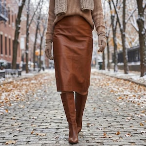 May include: A brown leather skirt paired with matching knee-high boots. The skirt is a midi length and the boots are a solid brown color. The outfit is completed with a tan sweater and scarf, creating a cohesive and stylish look.