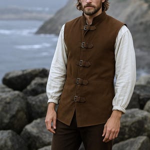 May include: A brown suede vest with four buckle closures, worn over a white long-sleeved shirt. The person is wearing a black tricorn hat and brown pants. The background shows a rocky shoreline and the ocean.