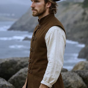 May include: A brown suede vest with buckle closures, worn over a white long-sleeve shirt. The outfit is completed with a black tricorn hat with a feather. The scene is set outdoors with a coastal backdrop.