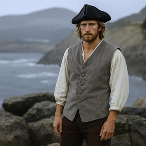 May include: A man wearing a black tricorn hat, gray vest with button closure, and a white long-sleeve shirt. The man is standing outdoors with a coastal background. The vest is a neutral gray color.
