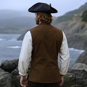 May include: A brown suede vest with a high collar, worn over a white long-sleeved shirt. A black tricorn hat with a feather is worn on the head. The outfit is reminiscent of historical or pirate-themed attire.