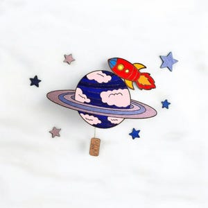 Può includere: Decorazione murale in legno a tema spaziale, raffigurante un pianeta blu con un anello, nuvole rosa e un razzo rosso. Stelle di legno di vari colori circondano il pianeta. Una piccola etichetta con testo è appesa sotto.