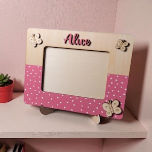 May include: Wooden picture frame with a pink polka dot design and the name "Alice" in pink script. The frame has three wooden butterfly embellishments. The frame is supported by a wooden stand.