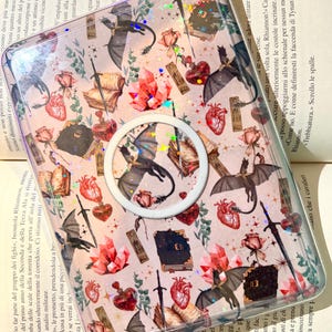 Può includere: Custodia trasparente per telefono con un design a tema fantasy. Presenta illustrazioni di draghi, cuori, libri, cristalli e spade. Include un pop-socket bianco. La custodia è decorata con glitter.