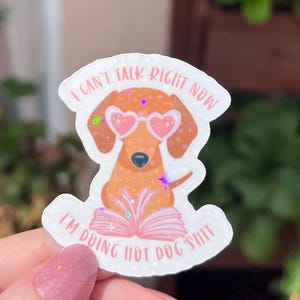 Könnte beinhalten: Ein Aufkleber mit einem Cartoon-Dackel, der eine herzförmige Sonnenbrille trägt. Der Text auf dem Aufkleber lautet: "I can't talk right now, I'm doing hot dog shit." Der Hund liest ein Buch. Der Aufkleber hat einen weißen Rand und holografische Akzente.