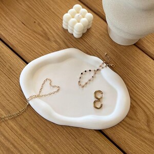 Könnte beinhalten: Ein weißes, organisch geformtes Keramiktablett mit Goldschmuck, darunter eine herzförmige Halskette und Creolen. Eine kleine, weiße Blasenkerze und eine dekorative Vase befinden sich ebenfalls auf der Holzoberfläche.