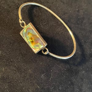 May include: A silver bangle bracelet with a rectangular abalone shell centerpiece. The shell displays iridescent colors of green, gold, and purple. The bracelet is on a dark background.