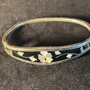 May include: Silver-tone hinged bangle bracelet with a black inlay. The inlay features a floral design with mother-of-pearl petals and small geometric accents. The bracelet is set against a dark background.