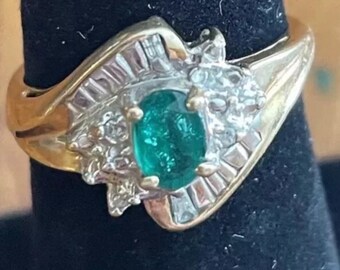 May include: A gold ring with a large emerald green gemstone surrounded by smaller diamonds. The ring has a unique, asymmetrical design.