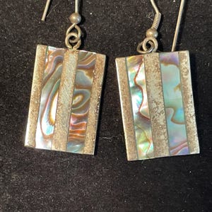 May include: A pair of rectangular earrings with iridescent abalone shell inlays. The earrings have a silver-tone metal frame and ear wires. The abalone shell displays a range of colors, including blue, green, and brown.