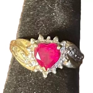 May include: A gold ring with a heart-shaped ruby gemstone surrounded by smaller diamonds. The ring has a twisted band with a diamond accent.