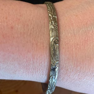May include: A metallic bangle bracelet with a textured surface featuring an intricate, raised floral and leaf pattern. The bracelet has a polished finish that catches the light, highlighting the detailed engravings across the entire outer band.