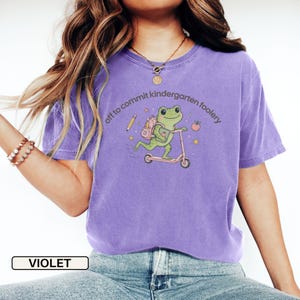 May include: A violet short-sleeve t-shirt featuring a graphic of a green frog riding a scooter with a backpack, a pencil, an apple, and stars. The text "off to commit kindergarten foolery" is printed in a curved arc above the frog illustration.