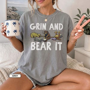 May include: A gray short-sleeve t-shirt featuring the white text "GRIN AND BEAR IT" above and below a graphic of three brown bears in various poses. One bear lies down, one sits with a book, and one lies on its back. A white mug with blue palm trees is held.
