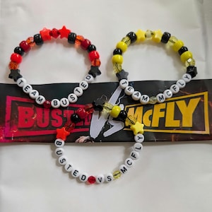 May include: Three beaded bracelets with the words "TEAM BUSTED" and "TEAM McFLY" spelled out in white letter beads. The bracelets feature red, black, and yellow beads, with star-shaped accents. The bracelets are on a background with the words "BUSTED" and "McFLY".