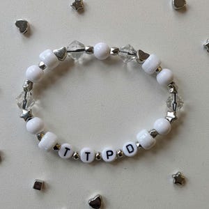 May include: White beaded bracelet with silver accents and letter beads spelling out "TTPD".