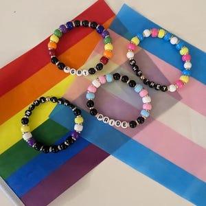 Op de afbeelding: Vier ronde kralenarmbanden met kleurrijke plastic kralen en witte letterkralen. Eén armband spelt LGBT, één spelt SHE THEY, één spelt HEY THEM en één spelt PRIDE. De armbanden zijn in een groepje op een plat oppervlak gerangschikt.