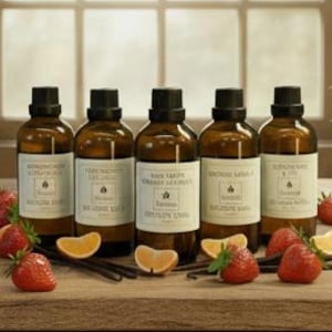 May include: Five amber glass bottles with black caps and white labels, arranged in a row. Each bottle features a central logo and text. Fresh strawberries and orange slices are placed in front of the bottles on a wooden surface.