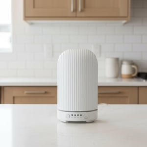Op de afbeelding: Een witte etherische olie diffuser met een geribbeld ontwerp. De diffuser heeft een afgeronde bovenkant en een basis met bedieningsknoppen. Hij staat op een wit aanrechtblad in een keuken. De achtergrond omvat witte metro tegels en houten kasten.