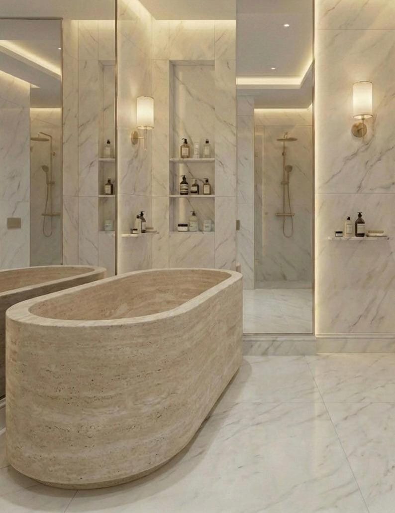 Puede incluir: Un lujoso ba&ntilde;o con una gran ba&ntilde;era de piedra beige como punto focal. Las paredes y el suelo est&aacute;n cubiertos de m&aacute;rmol blanco, y hay estantes empotrados con art&iacute;culos de tocador. El espacio est&aacute; iluminado por una iluminaci&oacute;n suave y apliques.