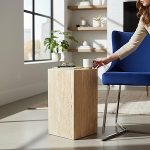 May include: A light beige, rectangular side table with a textured surface. A white mug with a beige base sits on top, next to a pair of glasses and a stack of books. A blue chair with silver legs is in the background.