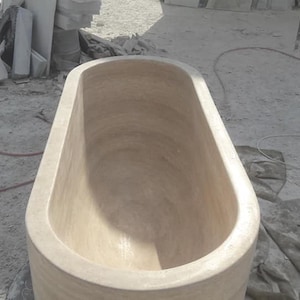 Puede incluir: Una gran ba&ntilde;era de forma ovalada, tallada en piedra de color claro. La ba&ntilde;era tiene bordes redondeados y un interior liso. La imagen est&aacute; tomada desde arriba, mostrando el interior de la ba&ntilde;era. El fondo incluye bloques de piedra y materiales de construcci&oacute;n.