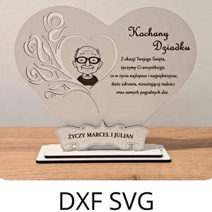 May include: A heart-shaped wooden plaque with a portrait of a man wearing glasses, and the words "Kochany Dziadku" and a message in Polish. The plaque sits on a white base with the text "Życzy Marcel i Julian".