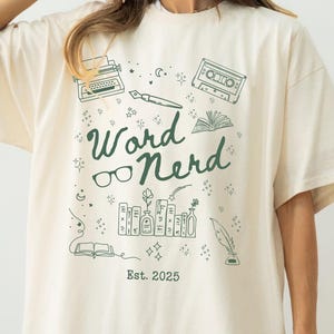 May include: Ivory-coloured t-shirt featuring a green "Word Nerd" graphic with illustrations of a typewriter, glasses, books, and a cassette tape. The text "Est. 2025" is also included. The shirt has a relaxed, oversized fit.