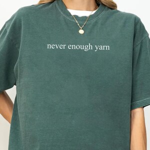 May include: A Blue Spruce coloured t-shirt with the white text "never enough yarn". The shirt is short-sleeved and has a crew neck. The t-shirt is a solid colour and has a relaxed fit.