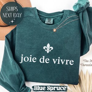 Puede incluir: Sudadera azul abeto con la frase "joie de vivre" en blanco, un s&iacute;mbolo de flor de lis y un delicado collar. El texto "Blue Spruce" est&aacute; en la parte inferior. La sudadera es de The Ivy Threads Co.