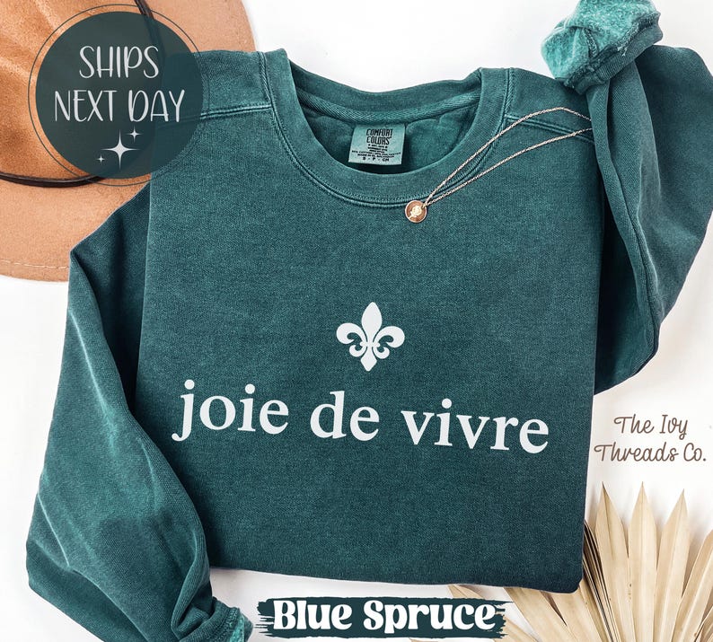 Puede incluir: Sudadera azul abeto con la frase "joie de vivre" en blanco, un s&iacute;mbolo de flor de lis y un delicado collar. El texto "Blue Spruce" est&aacute; en la parte inferior. La sudadera es de The Ivy Threads Co.