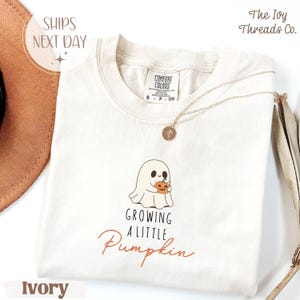 May include: Cream-coloured t-shirt with a round neck. It features a ghost holding a pumpkin graphic and the text "Growing a Little Pumpkin". A gold necklace is draped on the shirt. The shirt is labelled "Comfort Colors".
