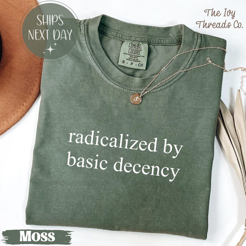 May include: A moss green t-shirt with the phrase "radicalized by basic decency" in white. The shirt has a crew neck and short sleeves. A gold necklace with a pendant is draped on the shirt. The text "Moss" is at the bottom.