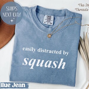 May include: A blue jean coloured t-shirt with the text "easily distracted by squash" in white. A gold necklace with a pendant rests on the shirt. The shirt is folded and has a Comfort Colors tag.