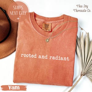 May include: A yam-colored t-shirt with the words "rooted and radiant" printed in white. The shirt has a crew neck and short sleeves. A gold necklace with a pendant rests on the shirt. The Ivy Threads Co. logo is visible.