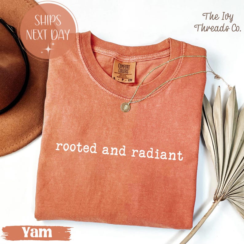 May include: A yam-colored t-shirt with the words "rooted and radiant" printed in white. The shirt has a crew neck and short sleeves. A gold necklace with a pendant rests on the shirt. The Ivy Threads Co. logo is visible.
