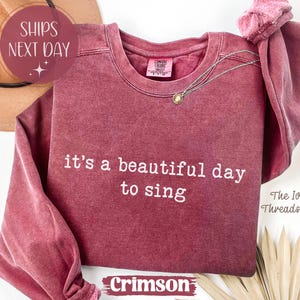 Op de afbeelding: Kleur kersenrood sweatshirt met de witte tekst "it's a beautiful day to sing". Het sweatshirt heeft lange mouwen en een ronde hals. Een gouden ketting is over de voorkant gedrapeerd.