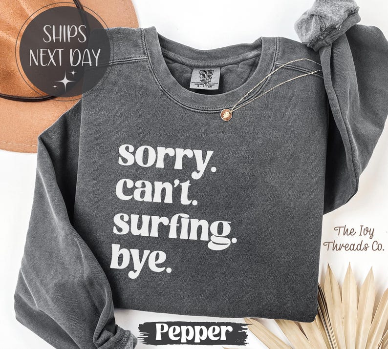 May include: A dark gray sweatshirt with the text "sorry. can't. surfing. bye." in white. The sweatshirt has long sleeves and a crew neck. A gold necklace is draped on the front. The word "Pepper" is at the bottom.