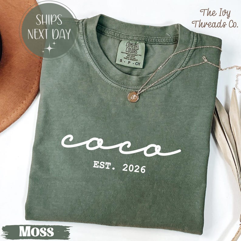 May include: A moss green t-shirt with the word "coco" in white script and "EST. 2026" printed below. The shirt has a crew neck and short sleeves. A gold necklace with a pendant is draped on the shirt. The Ivy Threads Co. label is visible.