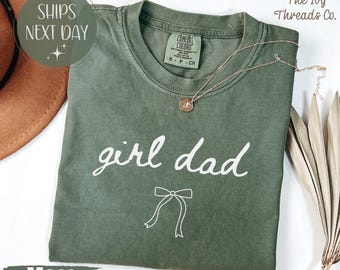 May include: Moss green t-shirt with the words "girl dad" in white script and a bow graphic. A gold necklace with a pendant rests on the shirt. The shirt is folded and displayed with a brown hat and dried plant.