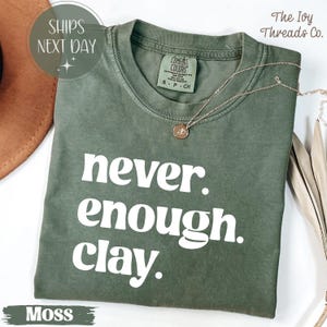 Puede incluir: Una camiseta verde musgo con el texto blanco "never enough clay". La camiseta tiene cuello redondo y mangas cortas. Un collar dorado con un colgante descansa sobre la camiseta. De The Ivy Threads Co.
