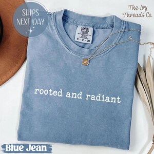 May include: A blue jean colored t-shirt with the words "rooted and radiant" in white. The shirt has a crew neck and short sleeves. A gold necklace with a pendant rests on the shirt. The tag reads "Comfort Colors".