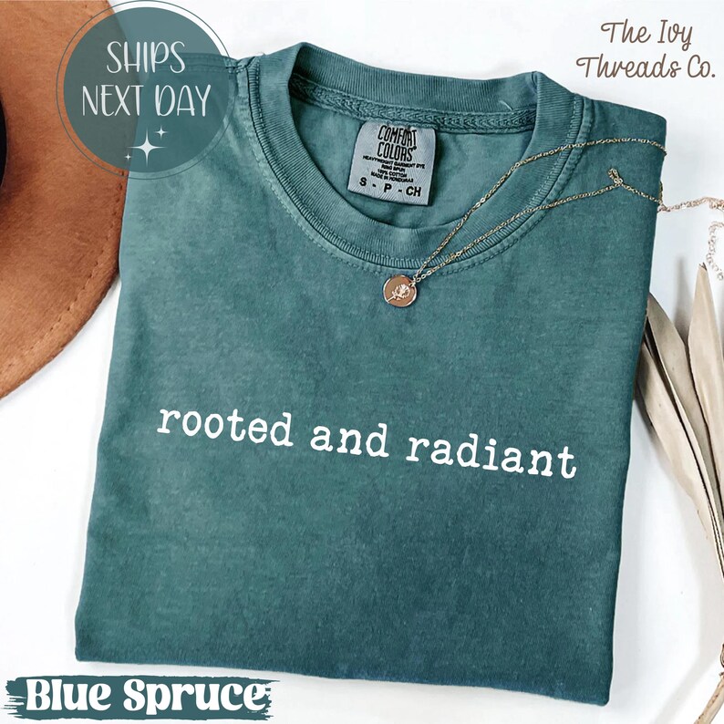 May include: A blue spruce t-shirt with the words "rooted and radiant" in white. The shirt has a crew neck and short sleeves. A gold necklace with a pendant is draped on the shirt. The text "SHIPS NEXT DAY" is in a circle.