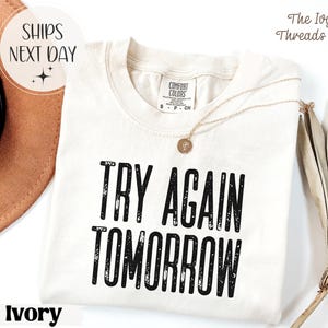 Könnte beinhalten: Elfenbeinfarbenes T-Shirt mit dem Text "TRY AGAIN TOMORROW" in fetten, schwarzen Buchstaben. Das Shirt hat einen Rundhalsausschnitt und kurze Ärmel. Eine goldene Halskette mit Anhänger liegt auf dem Shirt. Das Logo von The Ivy Threads Co. ist sichtbar.