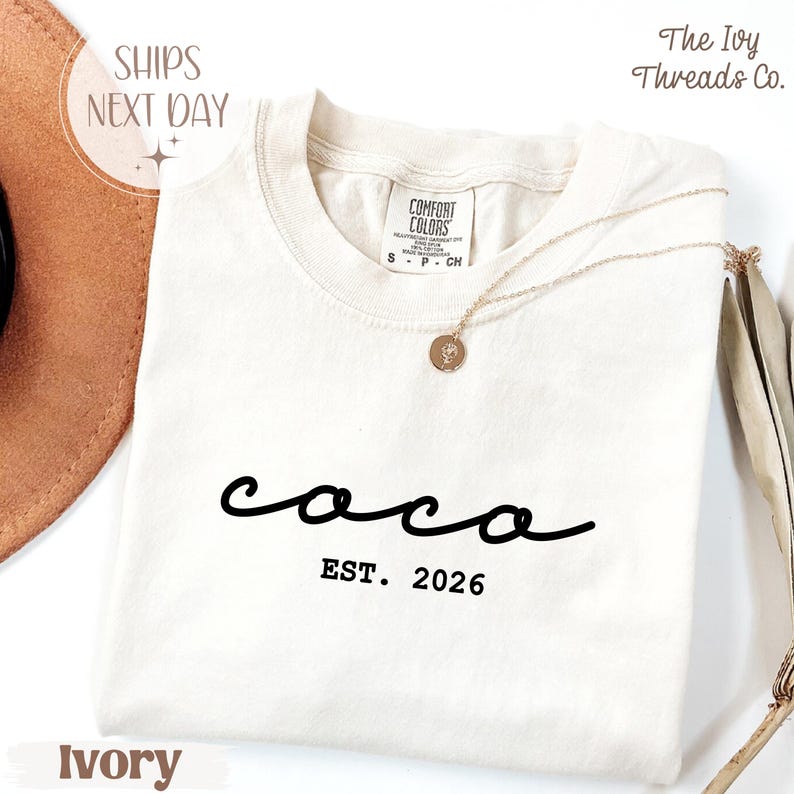 May include: Ivory-colored t-shirt with the word "coco" in a cursive font and "EST. 2026" printed below. The shirt has a crew neck and a small tag on the upper back. A gold necklace with a pendant rests on the shirt.