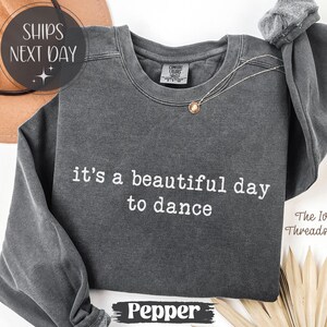 Puede incluir: Una sudadera gris oscuro con la frase "it's a beautiful day to dance" en letras blancas. La sudadera de manga larga tiene cuello redondo y un collar dorado. La palabra "Pepper" está impresa en la parte inferior. El logo de The Ivy Threads Co. es visible.