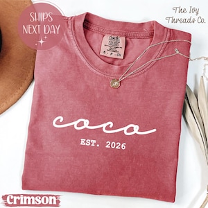 May include: Crimson-colored t-shirt with the word "coco" in white script and "EST. 2026" printed below. A silver necklace with a pendant rests on the shirt. The shirt is from The Ivy Threads Co.
