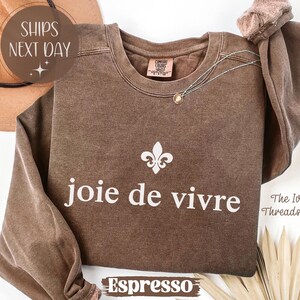 Puede incluir: Sudadera color espresso con la frase "joie de vivre" en blanco, junto con un s&iacute;mbolo de flor de lis. La sudadera tiene cuello redondo y mangas largas. Un collar dorado est&aacute; drapeado en la parte delantera.
