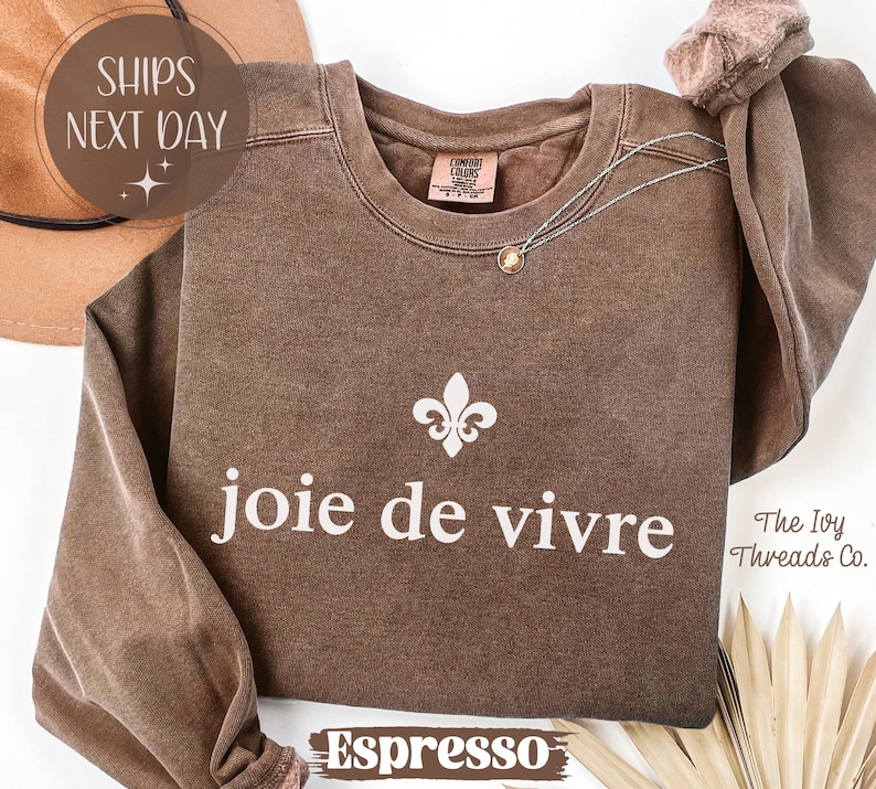 Puede incluir: Sudadera color espresso con la frase "joie de vivre" en blanco, junto con un s&iacute;mbolo de flor de lis. La sudadera tiene cuello redondo y mangas largas. Un collar dorado est&aacute; drapeado en la parte delantera.