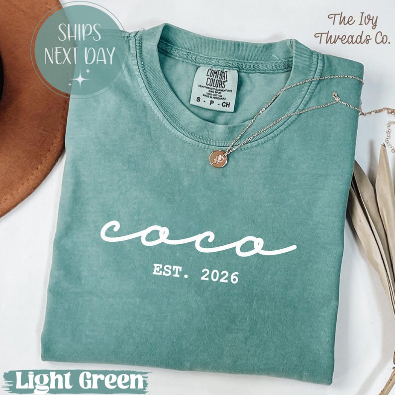 May include: A light green t-shirt with the word "coco" in white script and "EST. 2026" below it. A silver necklace with a pendant rests on the shirt. The shirt is folded and has a tag that says "Comfort Colors".